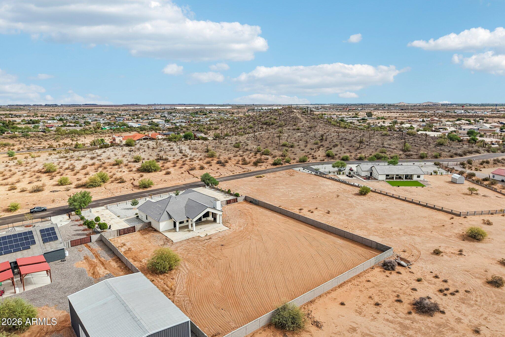 Aerial view of a desert home with a large fenced, graded yard and detached outbuildings.