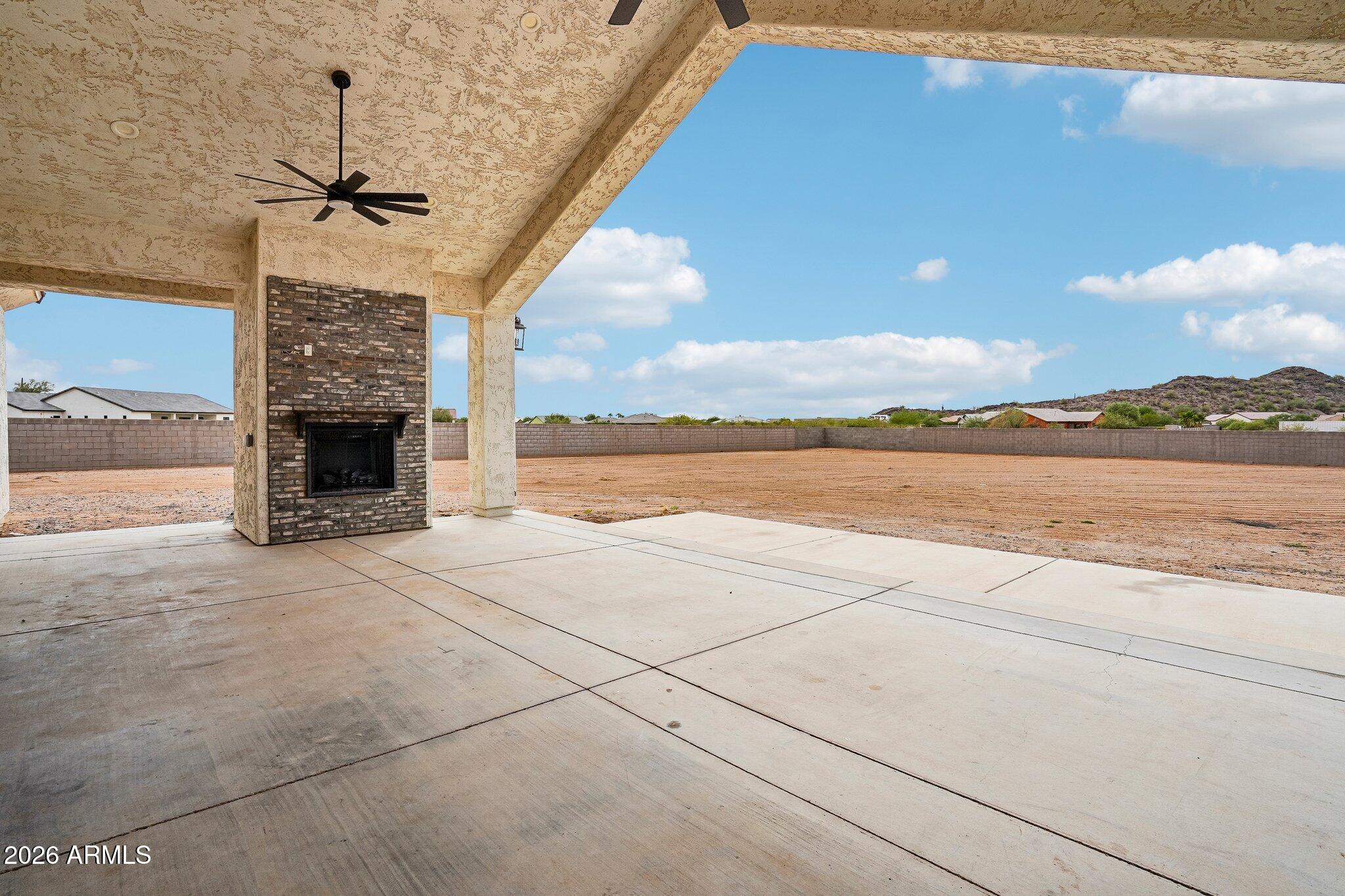 Covered concrete patio with a brick fireplace overlooks a large bare dirt yard and low desert hills.