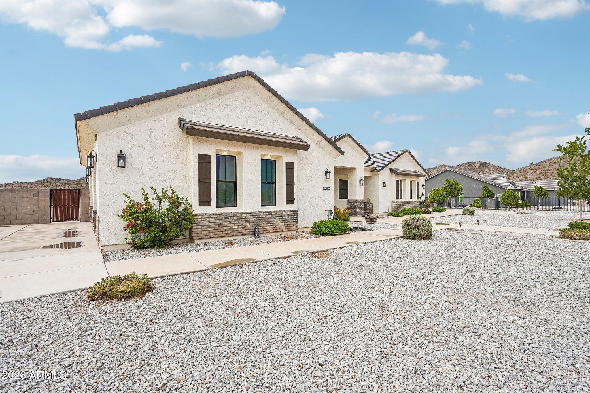 Single-story stucco home with a dark tile roof, gravel landscaping, and low desert hills beyond.