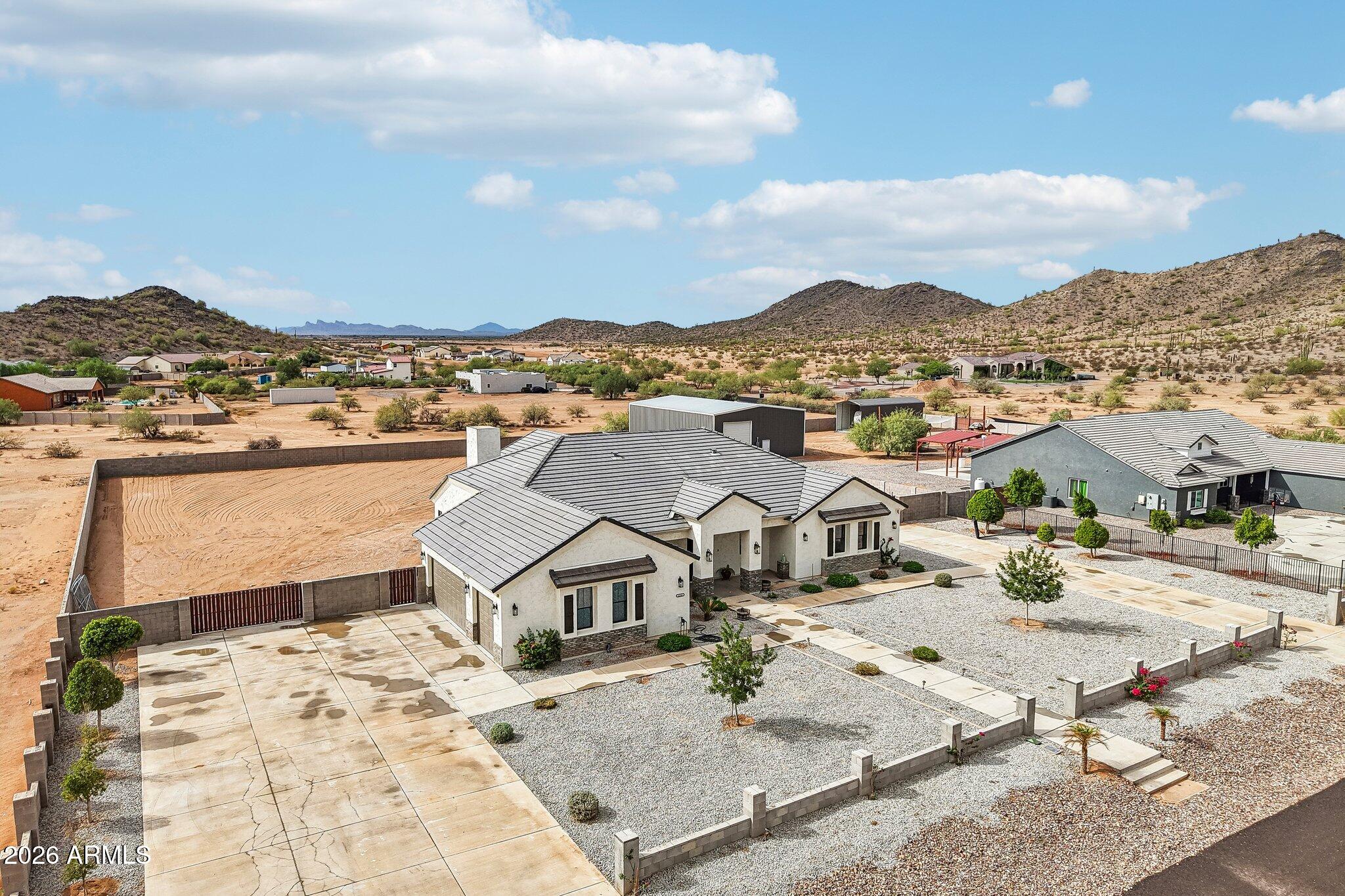 Aerial view of a stucco home with a broad driveway, gravel yard, and detached metal outbuilding among desert hills.