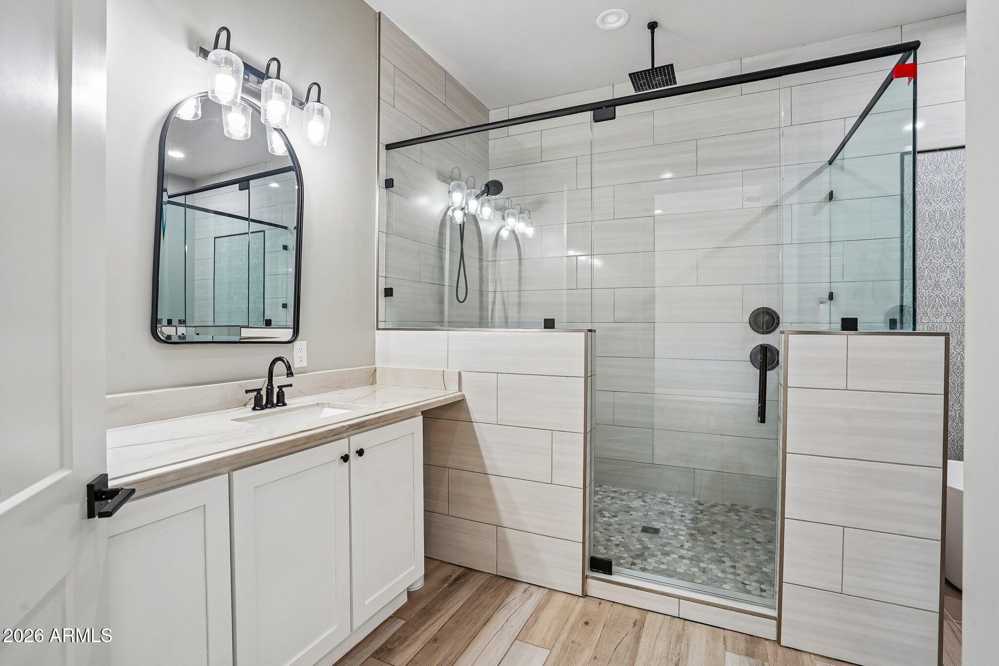 Bathroom with white vanity and arched mirror beside a glass walk-in shower with pale tile and black fixtures.