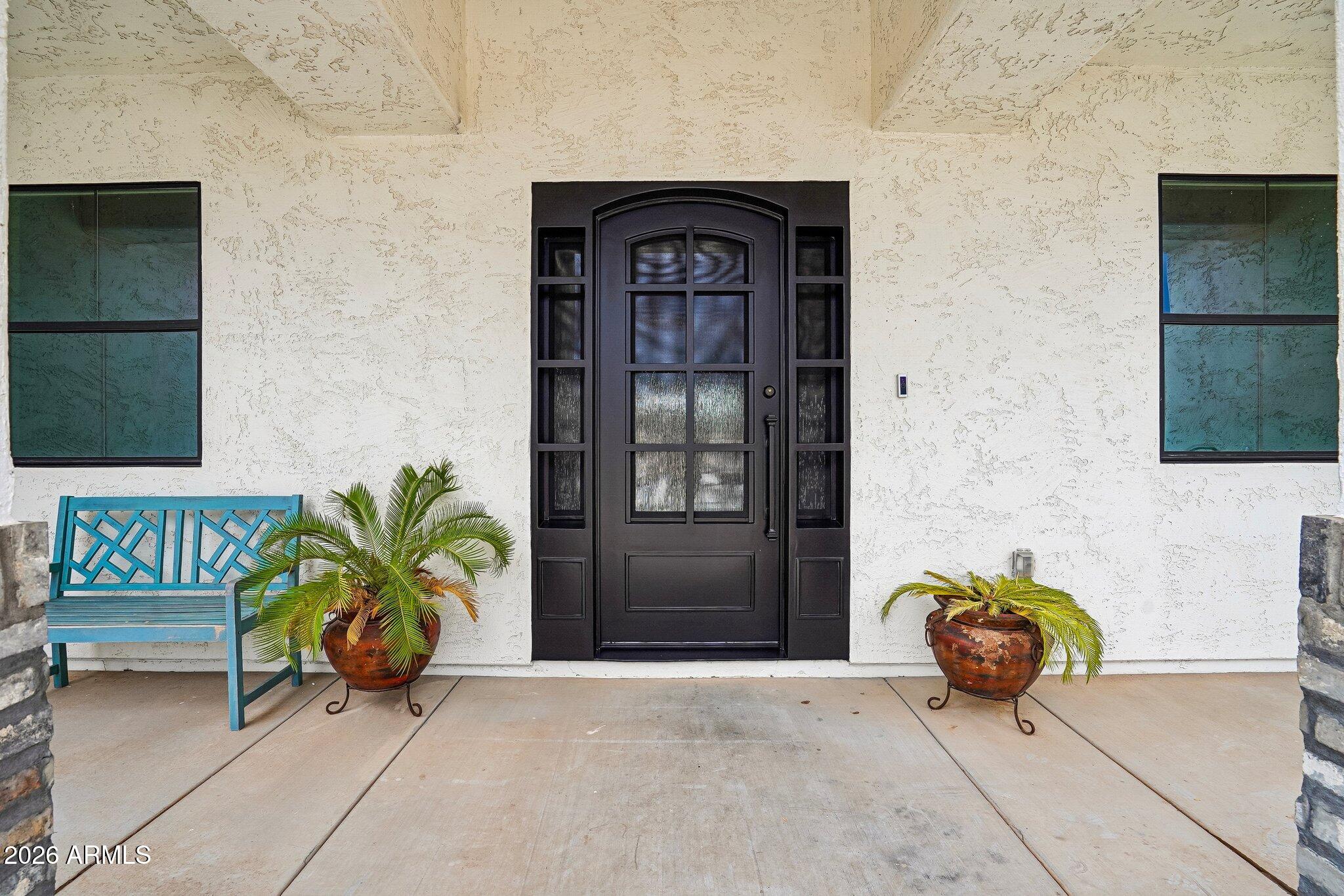 Home’s covered front entry with an arched dark door, turquoise bench, and potted palms.