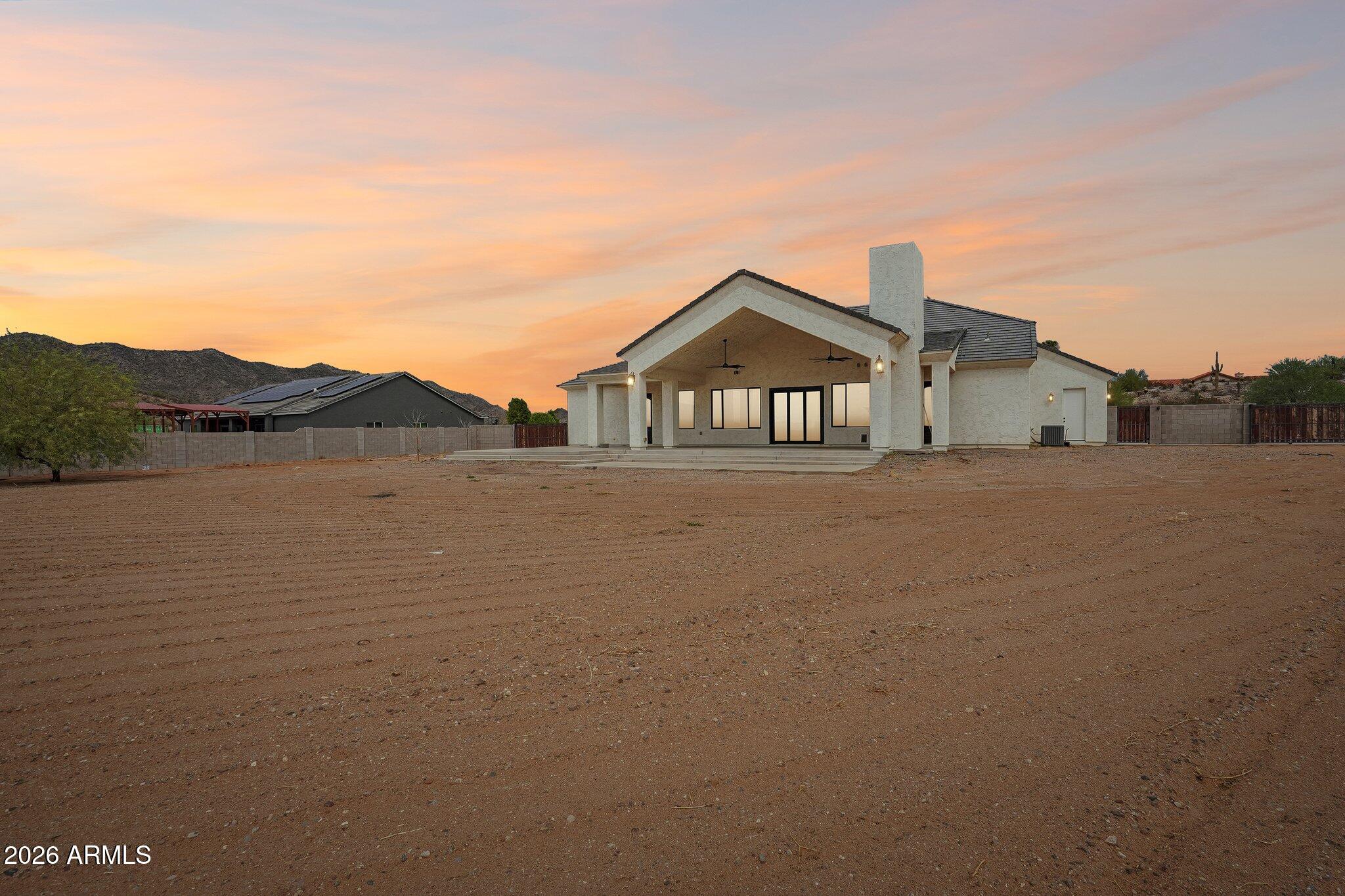 Home’s rear exterior with a covered patio, broad dirt yard, and desert hills at dusk.