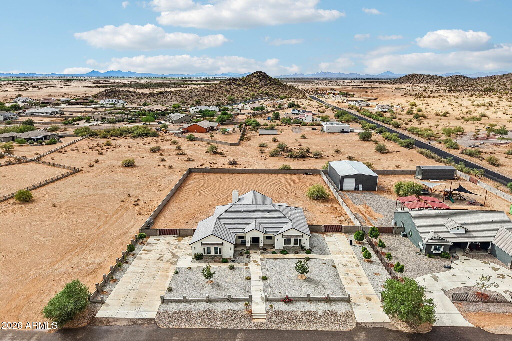 Aerial view of a desert home with a fenced lot, detached metal outbuilding, and distant mountains.