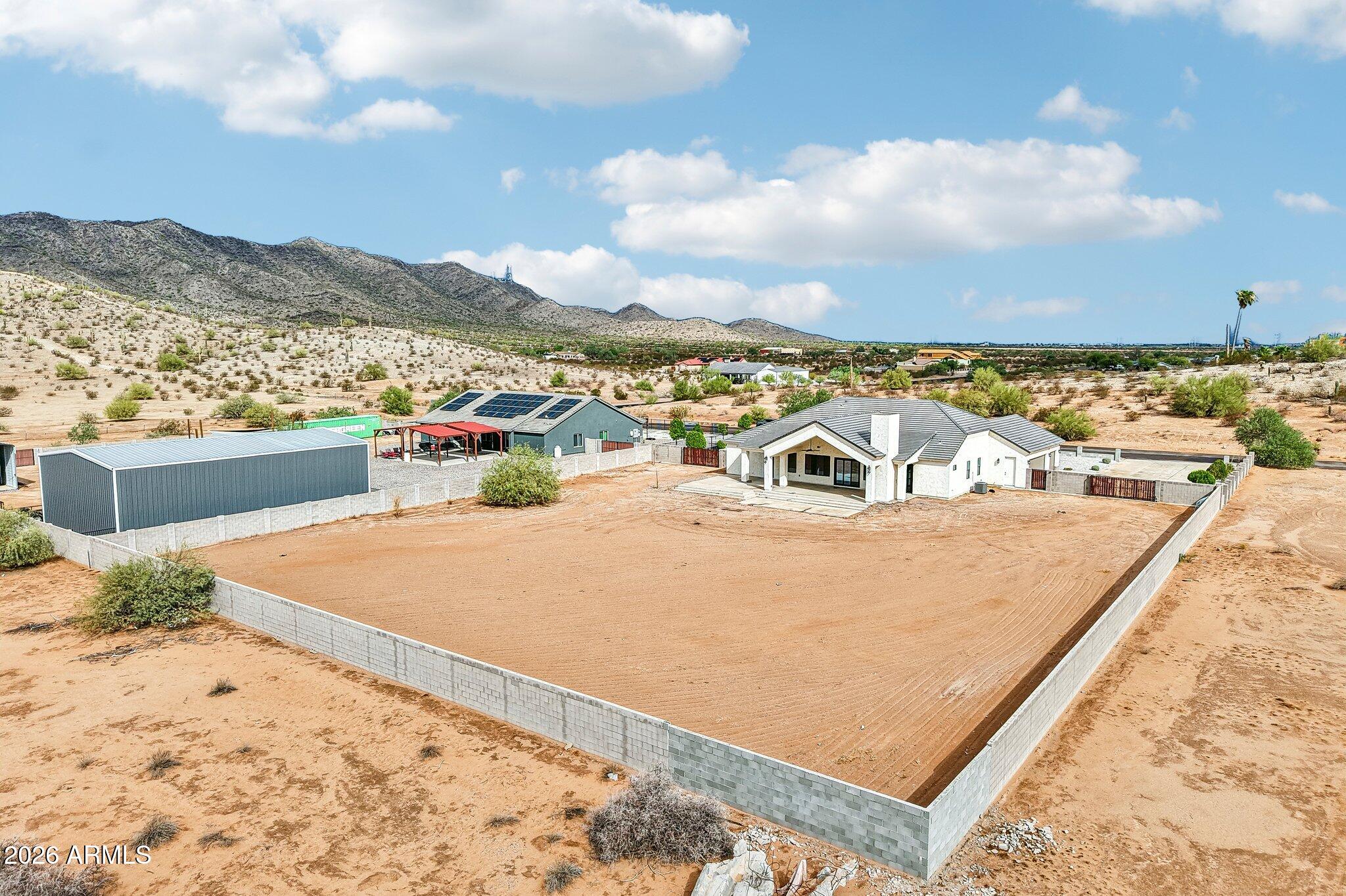 Aerial view of a white desert home, fenced dirt yard, solar-paneled outbuilding, and mountain backdrop.
