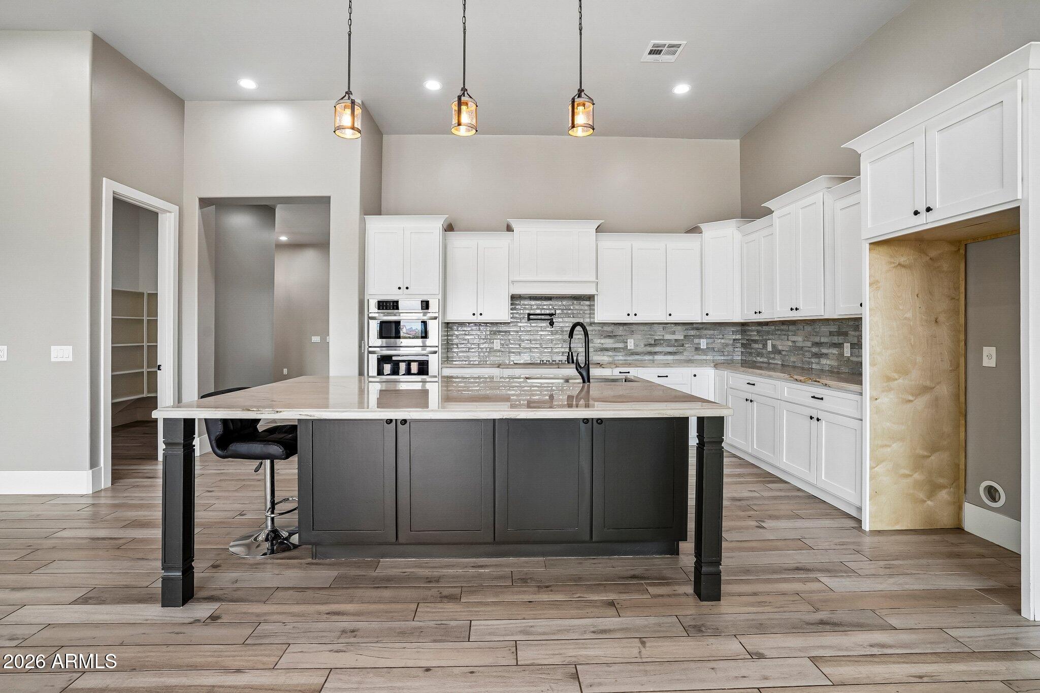 Kitchen with a large dark island, white cabinetry, gray tile backsplash, and double wall ovens.