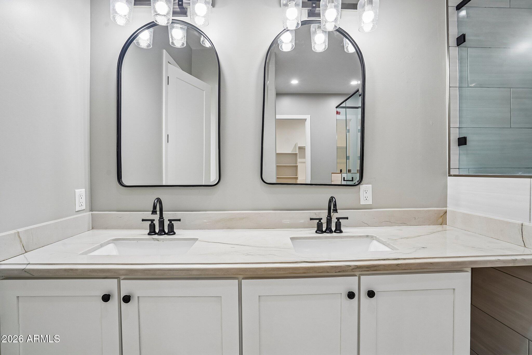 Bathroom vanity with double sinks, black faucets, arched mirrors, and glass shower doors.