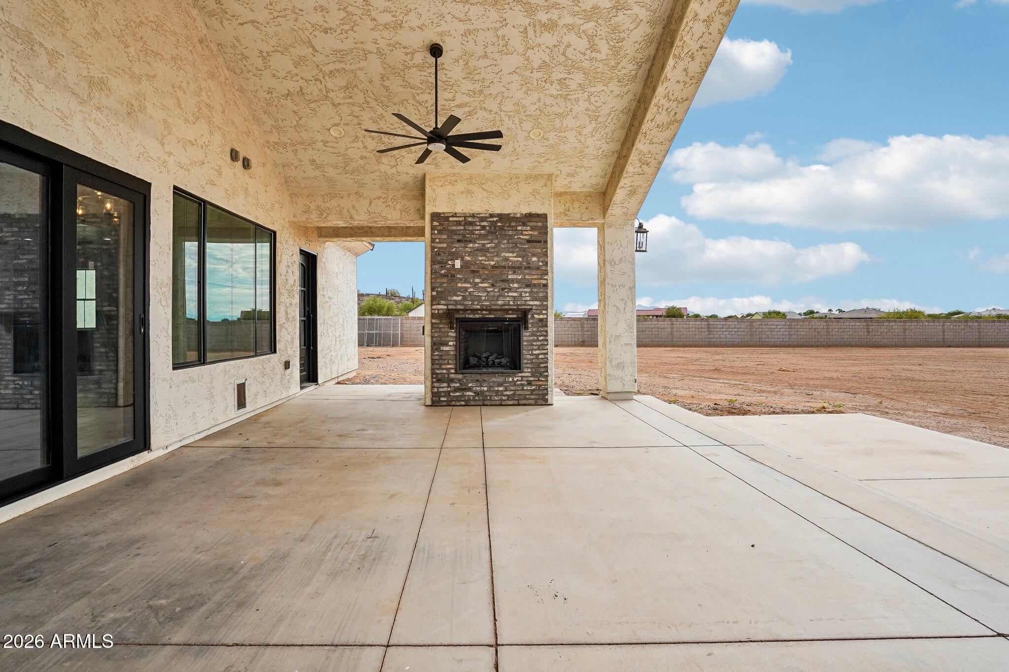 Covered patio with a brick fireplace and ceiling fan overlooking a fenced, bare yard.