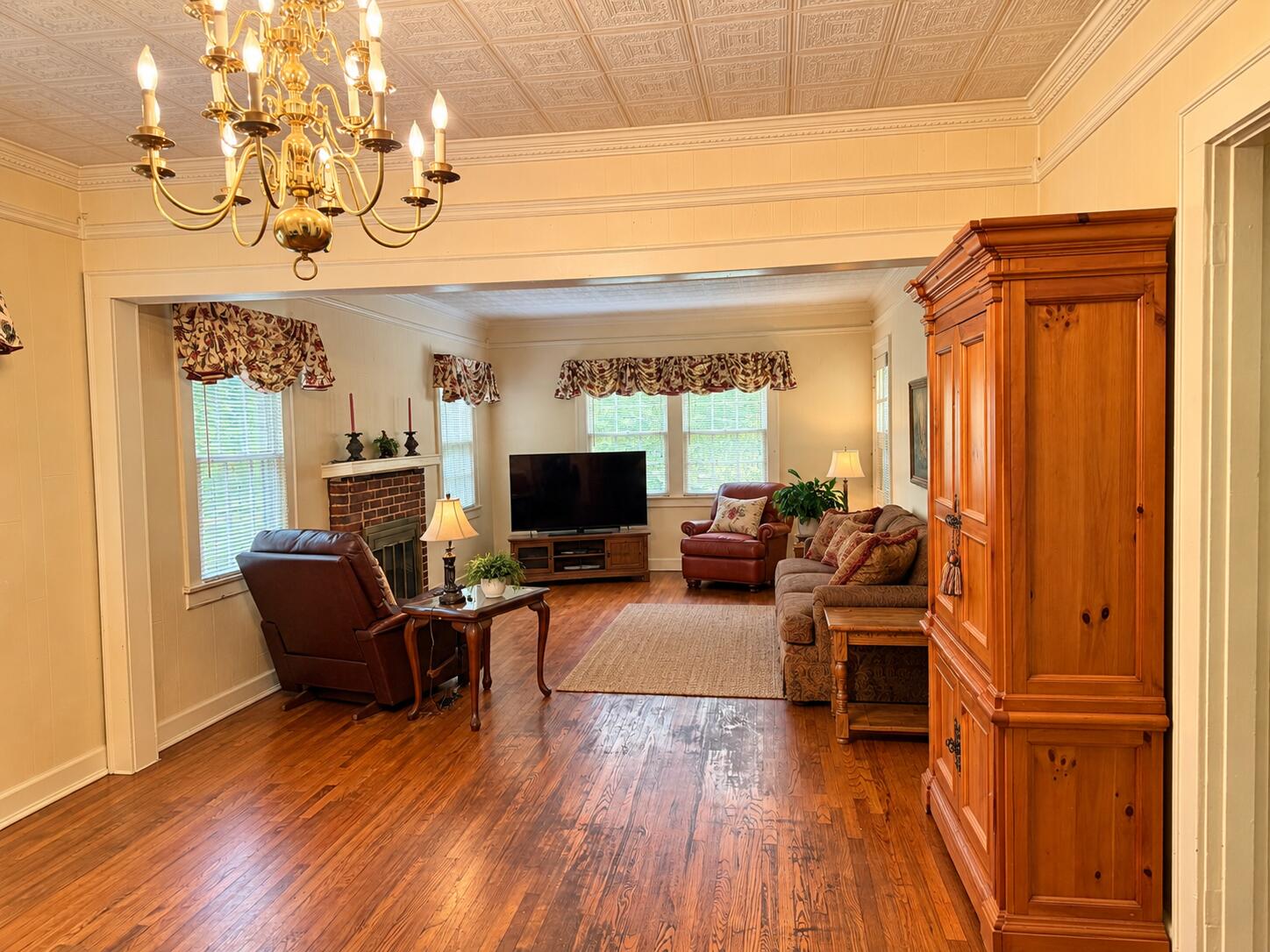 Living room with hardwood floors, brick fireplace, ornate ceiling, and brass chandelier.