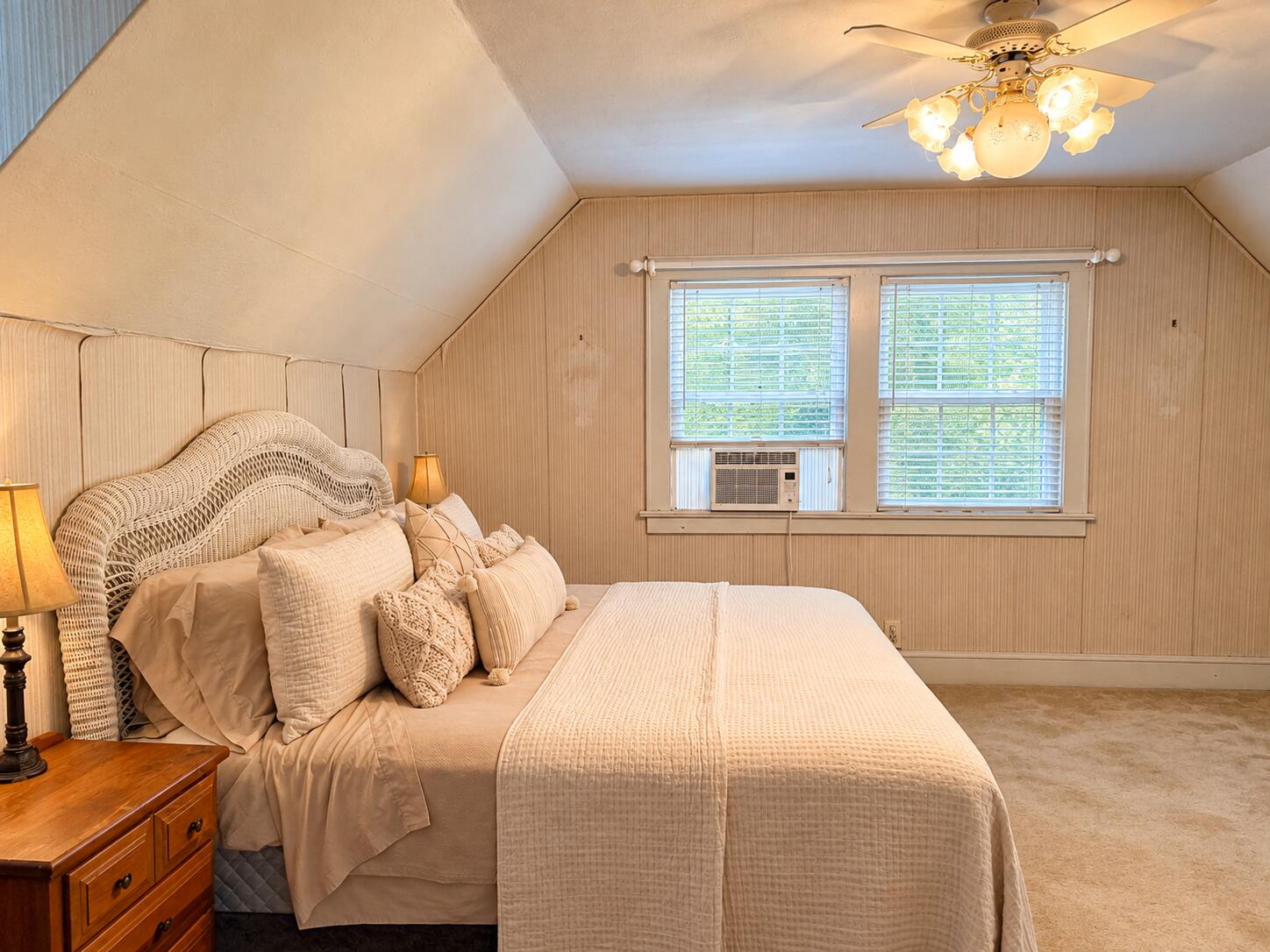 Bedroom with a woven wicker headboard, beige bedding, sloped ceiling, and two windows with a window air conditioner.