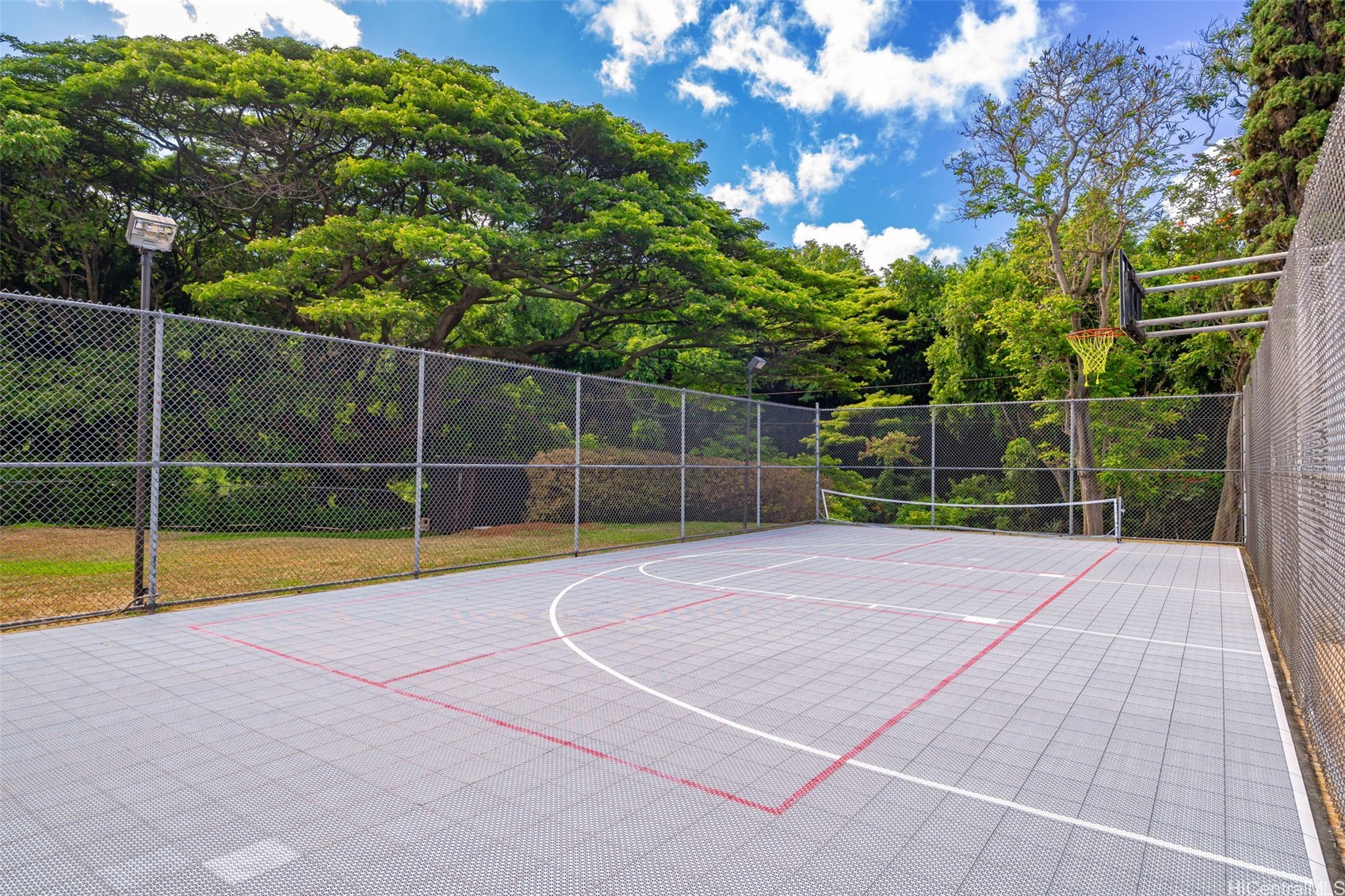 Outdoor basketball court with a gray tiled surface, painted lines, hoop, and chain-link fencing.