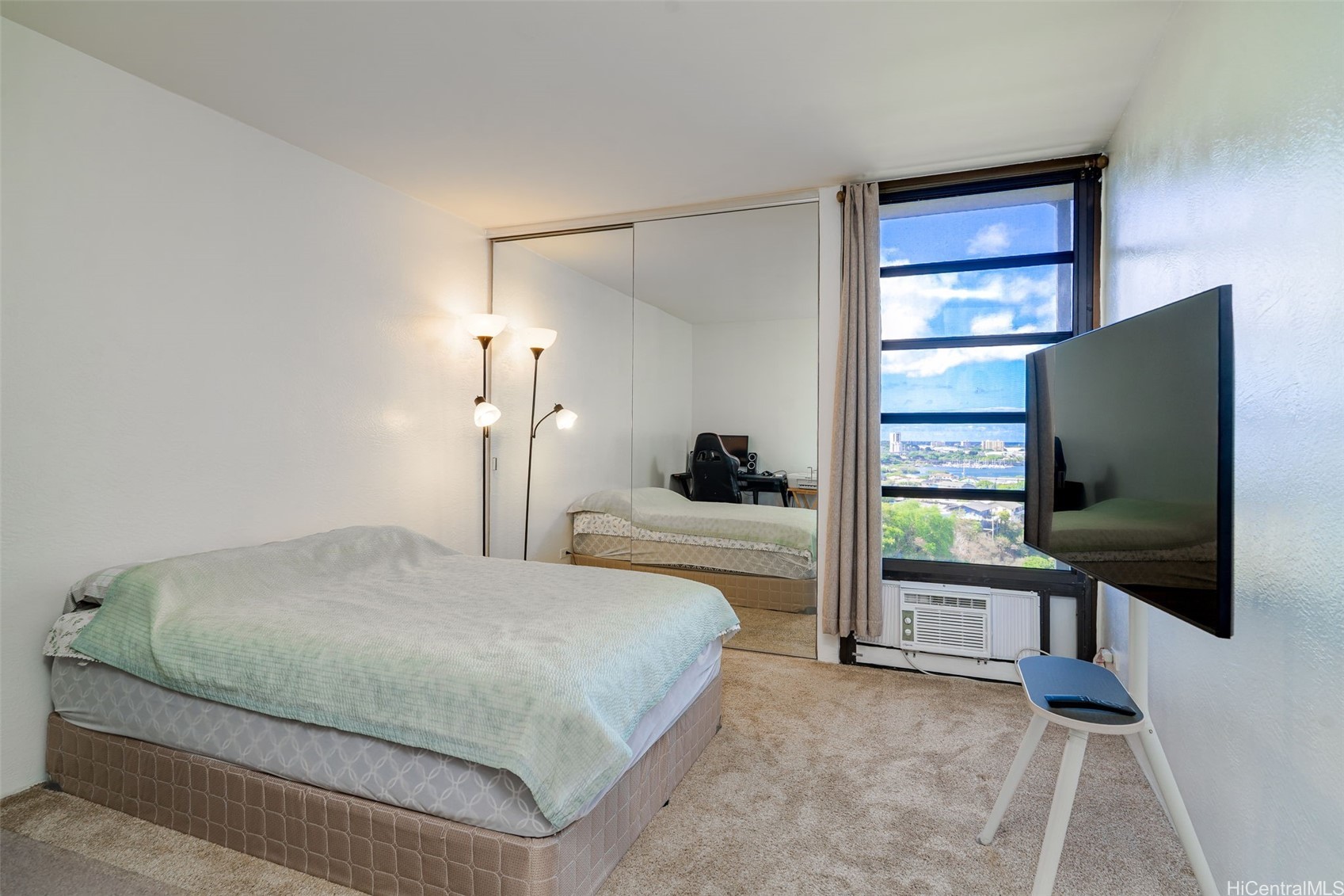 Bedroom with mirrored closet doors, a wall-mounted TV, and a view of water and distant buildings.