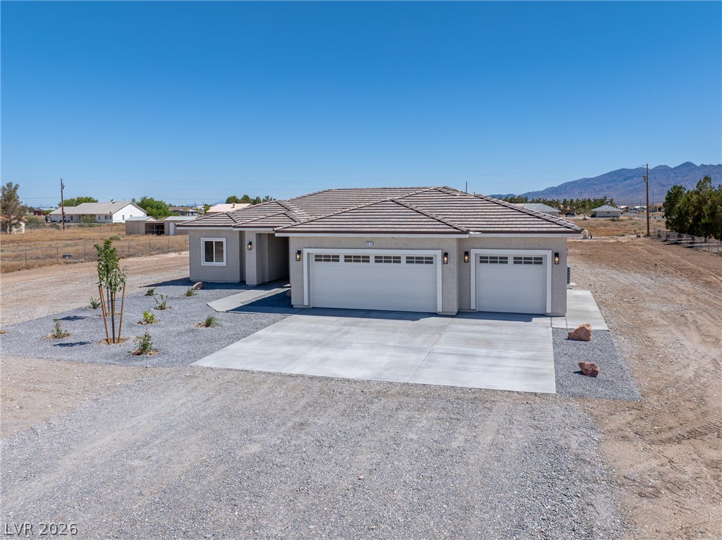 Single-story gray stucco home with a three-car garage, gravel lot, and distant mountain backdrop.