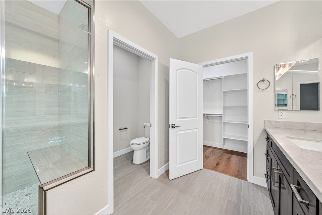 Modern bathroom with glass shower, separate toilet alcove, dark vanity, and walk-in closet.