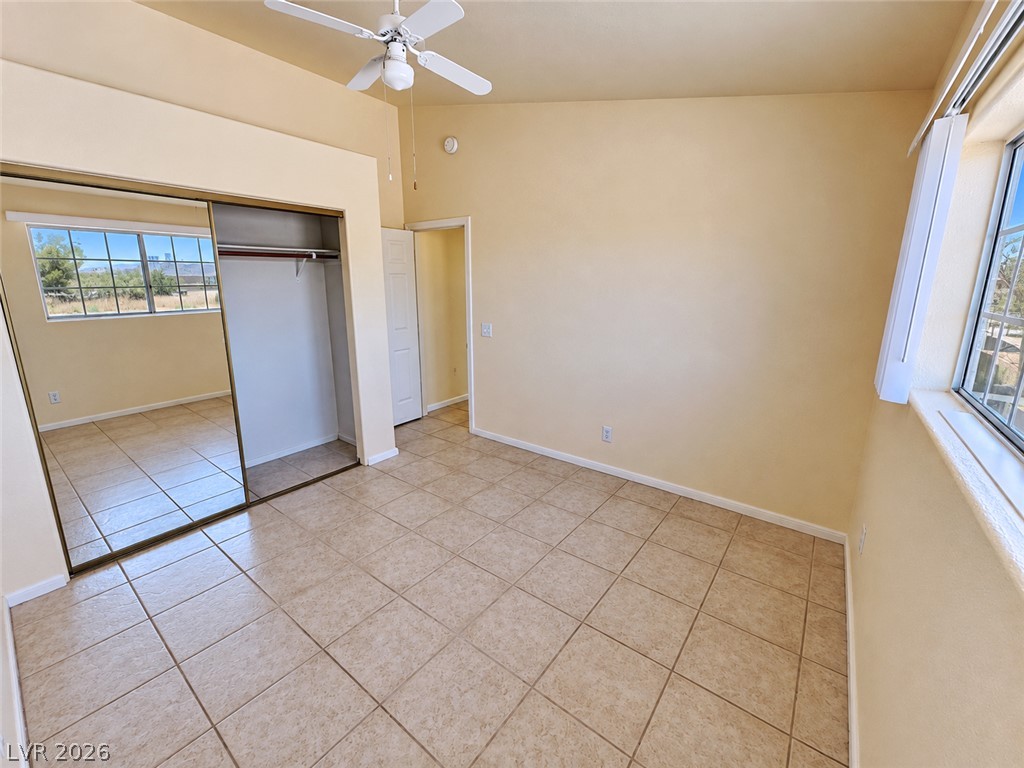 Empty bedroom with tan tile floors, mirrored closet doors, ceiling fan, and two windows.