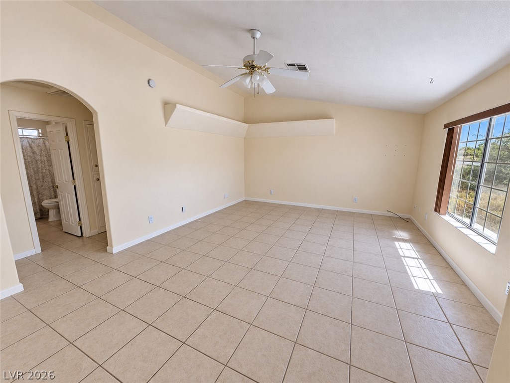 Empty tiled room with a ceiling fan, large window, and doorway to a bathroom.