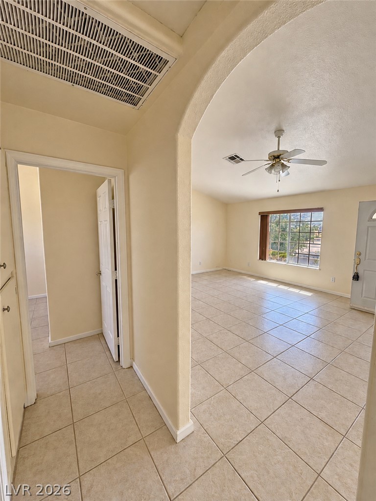 Vacant tiled living room with an arched hallway opening, ceiling fan, and window with vertical blinds.