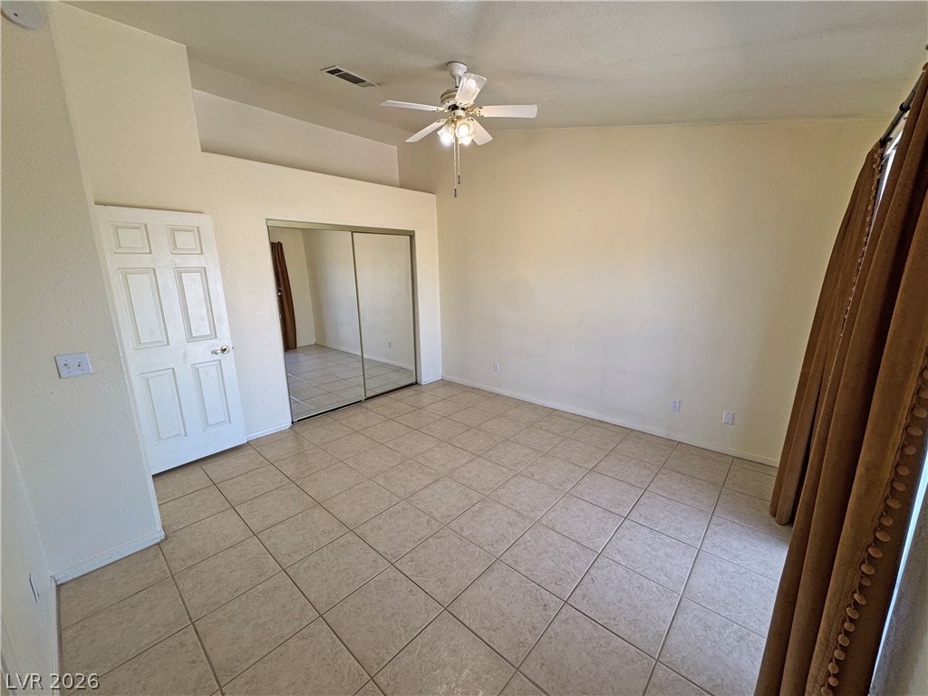 Empty bedroom with beige tile floors, mirrored closet doors, and a ceiling fan.