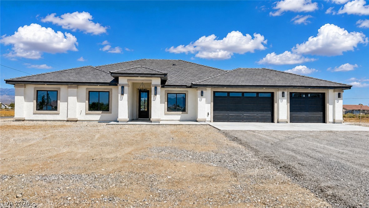 Single-story stucco home with a gray tile roof, two garage doors, and a broad gravel yard.