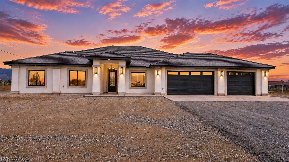 Single-story stucco home with two garage doors and a gravel yard at sunset.