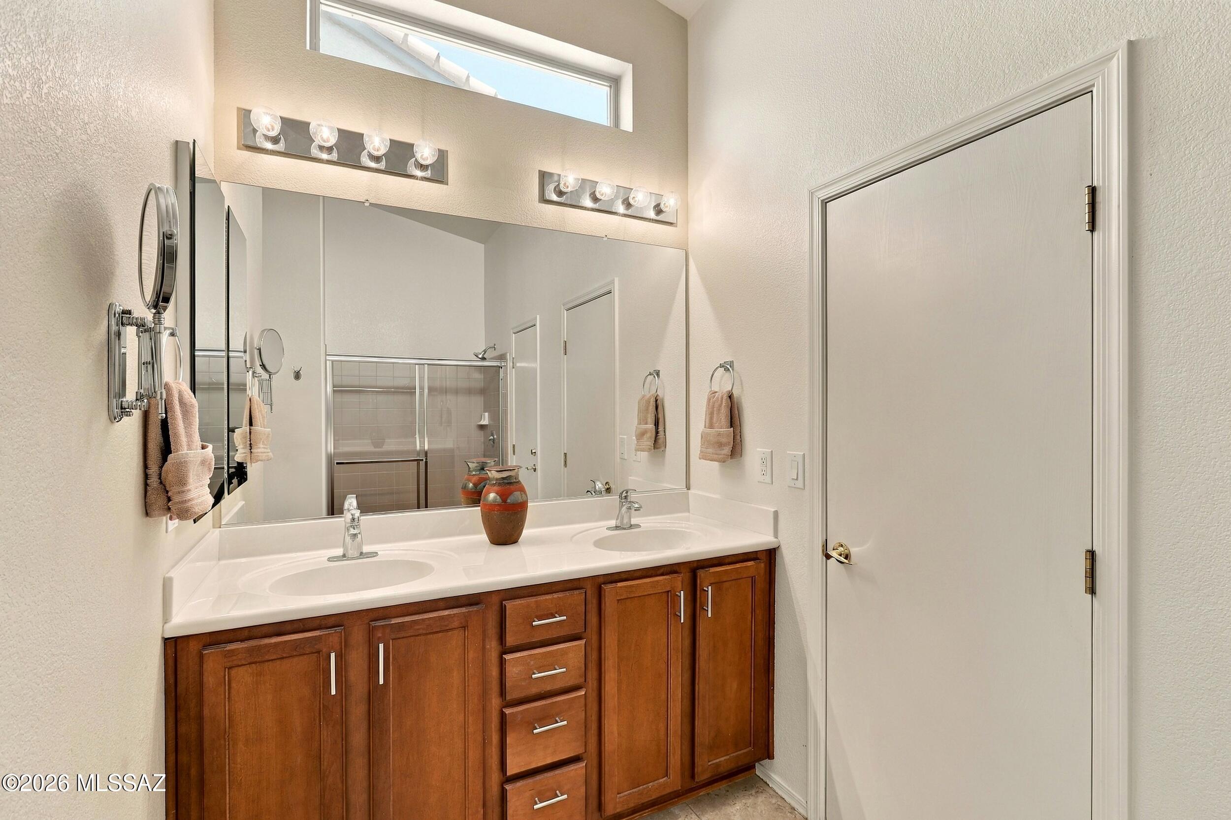 Bathroom with a double-sink vanity, warm wood cabinetry, and a tiled shower reflected in the large mirror.