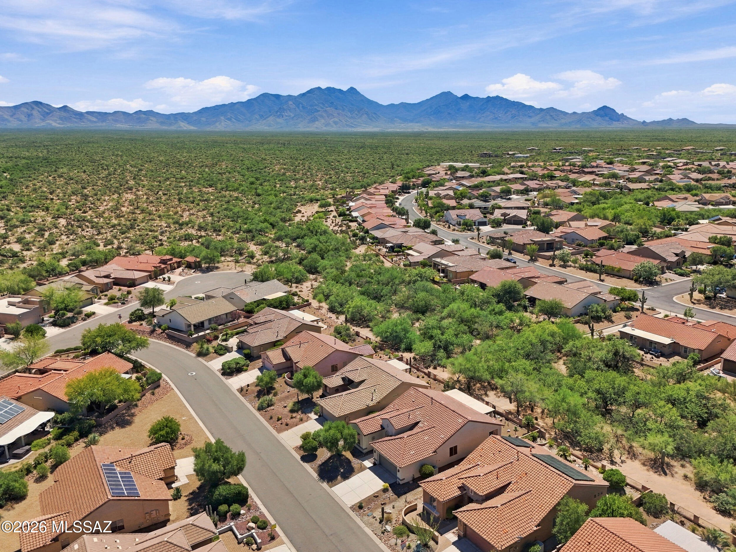 Aerial neighborhood view with tile-roof homes, open desert, and mountains in the distance.