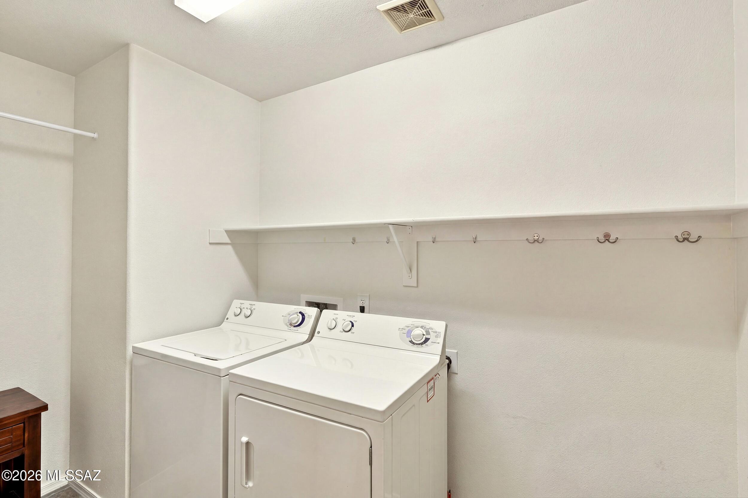 Laundry room with white washer and dryer beneath a long shelf with wall hooks.