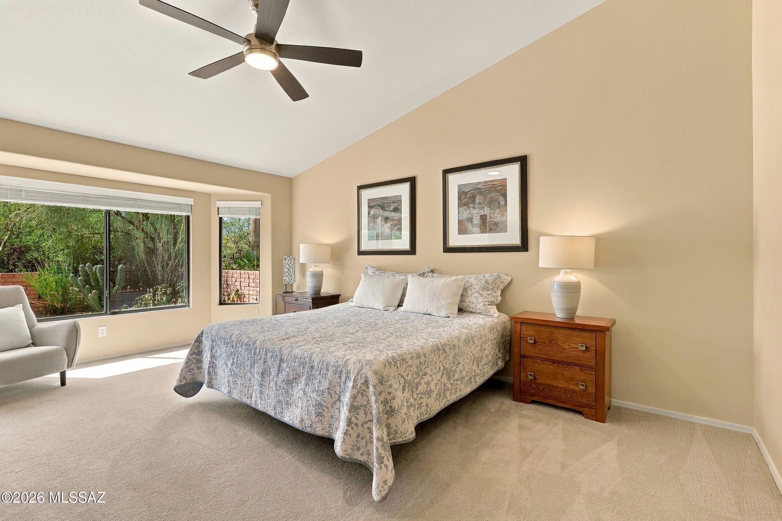 Furnished bedroom with a floral-covered bed, ceiling fan, and windows overlooking cacti and greenery.
