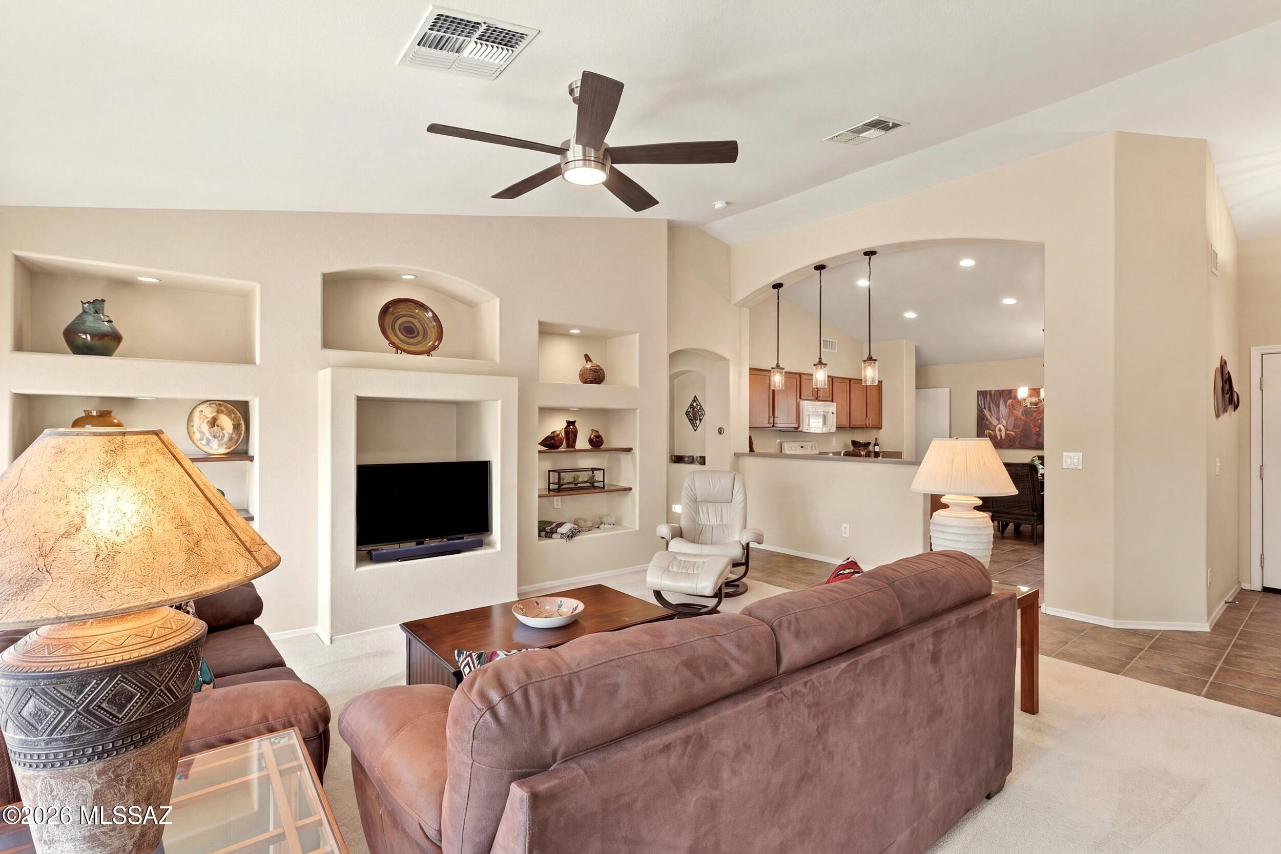 Living room with built-in TV niche and display shelves, brown sectional, and an open view into the kitchen.