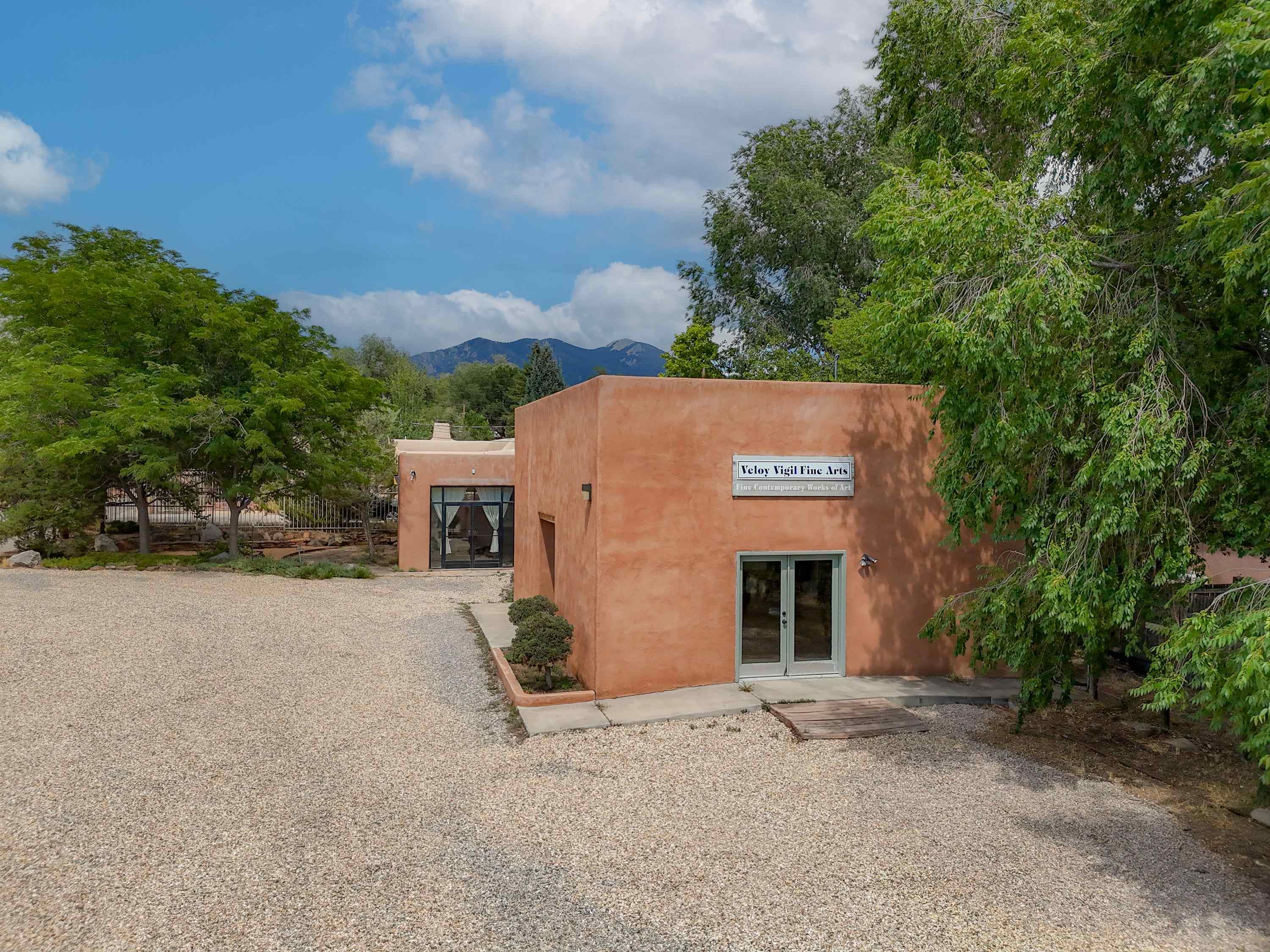 Adobe art gallery with a gravel courtyard, mature trees, and mountains in the background.