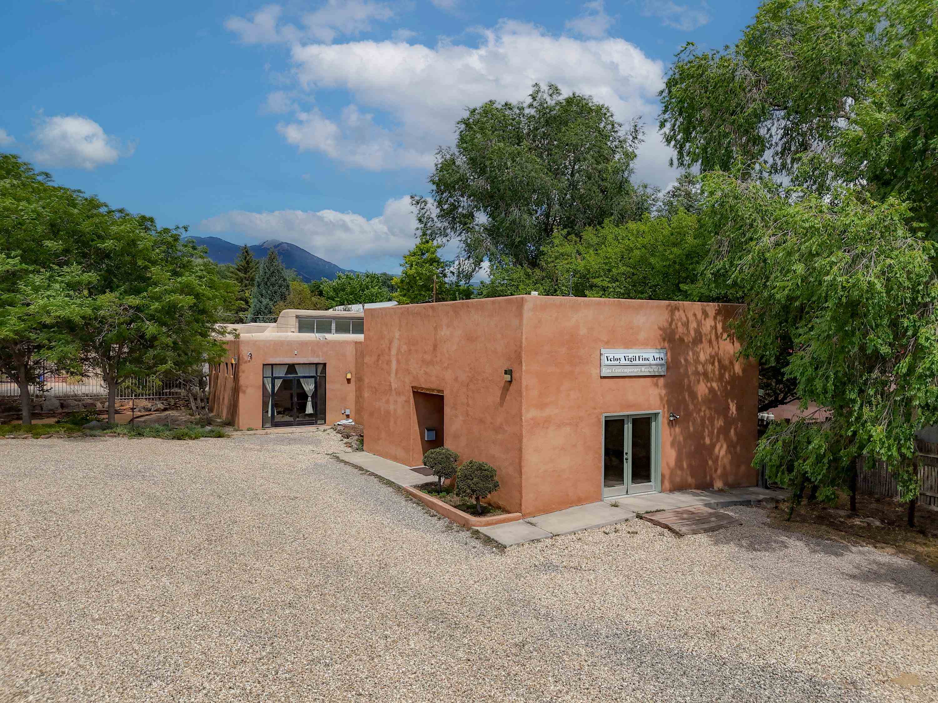 Adobe-style building with a gravel forecourt, leafy trees, and mountains behind it.