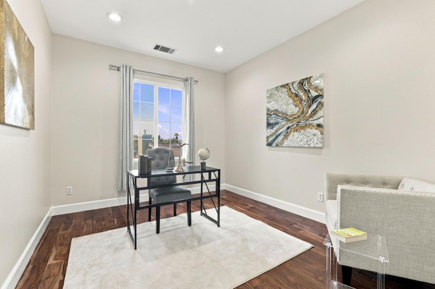 Staged home office with a black desk, curtained window, dark wood flooring, and light area rug.