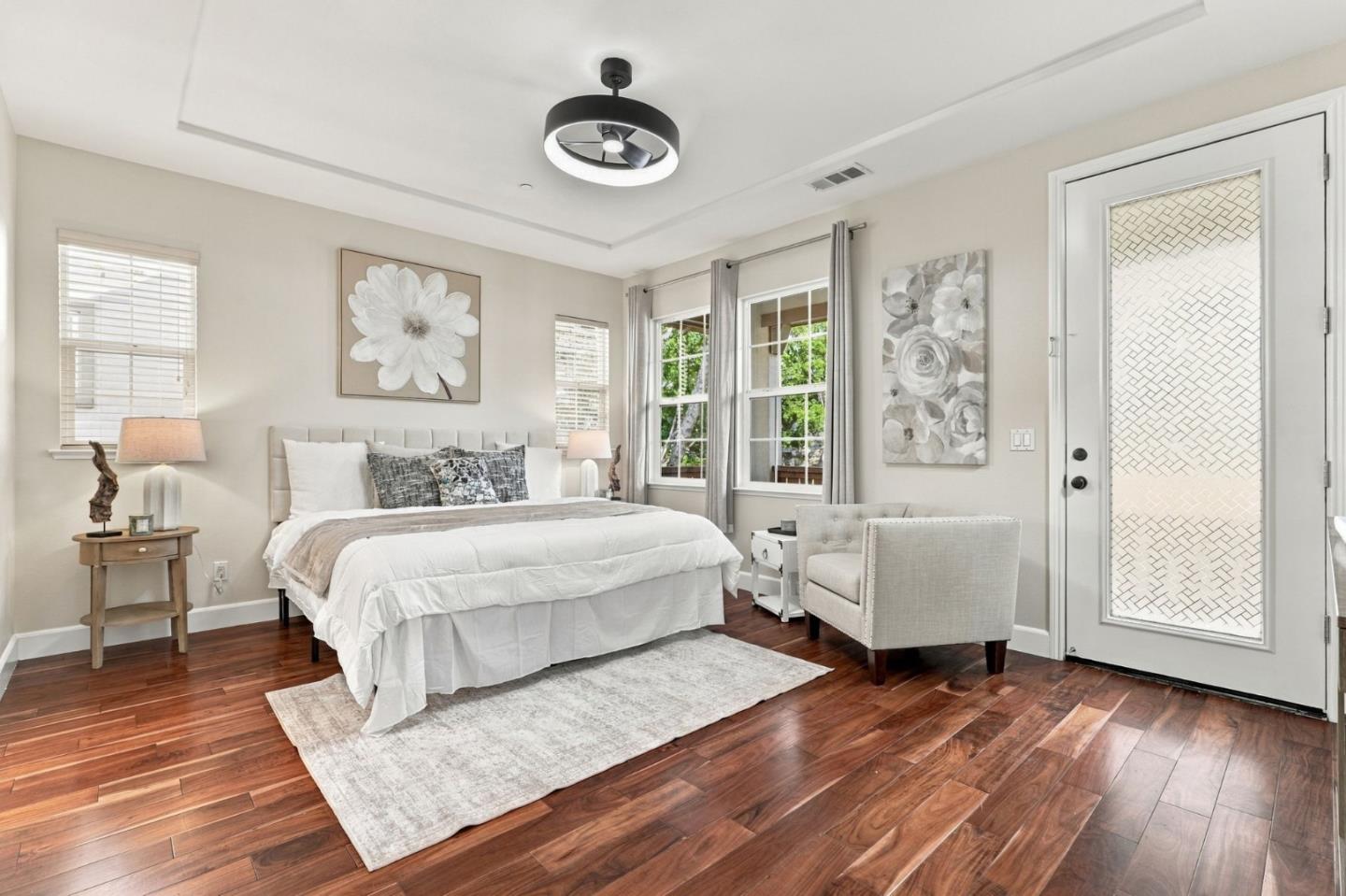 Staged bedroom with a large bed, dark wood floors, floral art, and windows overlooking greenery.