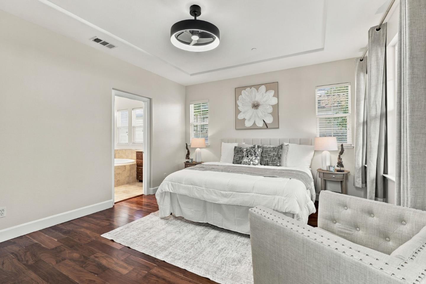 Staged bedroom with a white bed, dark wood floors, tray ceiling, and adjoining bathroom doorway.