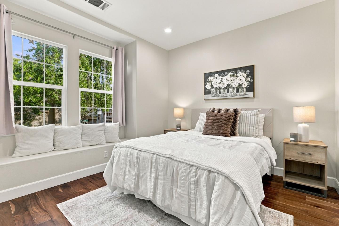 Staged bedroom with a white bed, window seat, and two large windows overlooking trees.