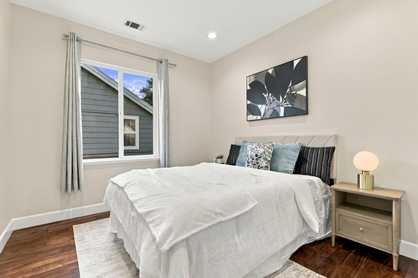 Staged bedroom with a white bed, blue accent pillows, botanical artwork, and dark wood floors.