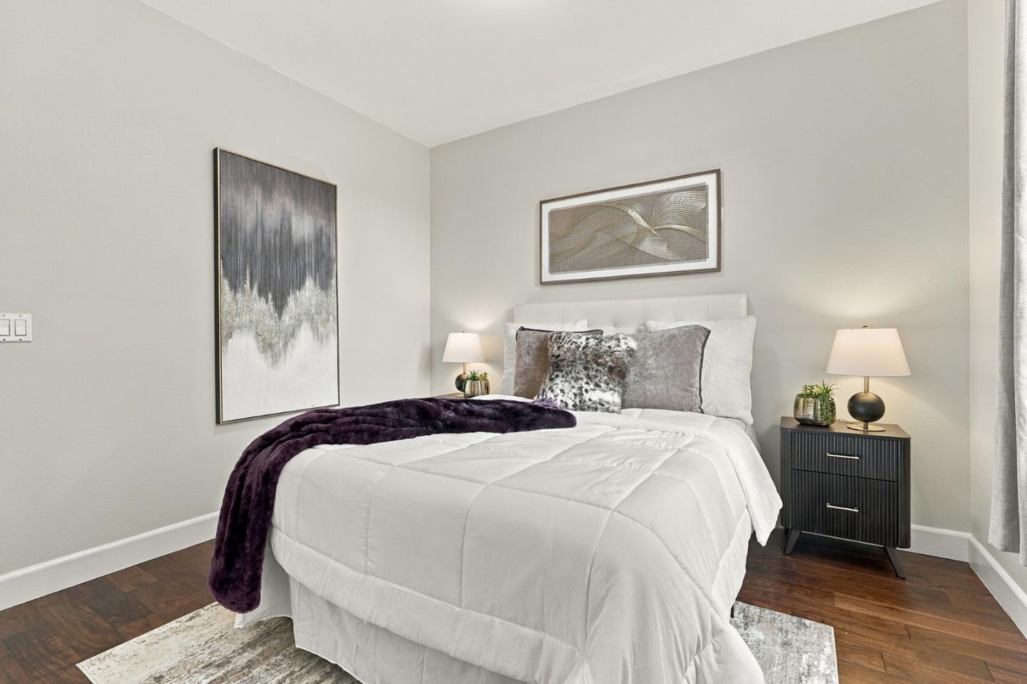 Staged bedroom with a white bed, deep purple throw, dark nightstand, and hardwood floors.
