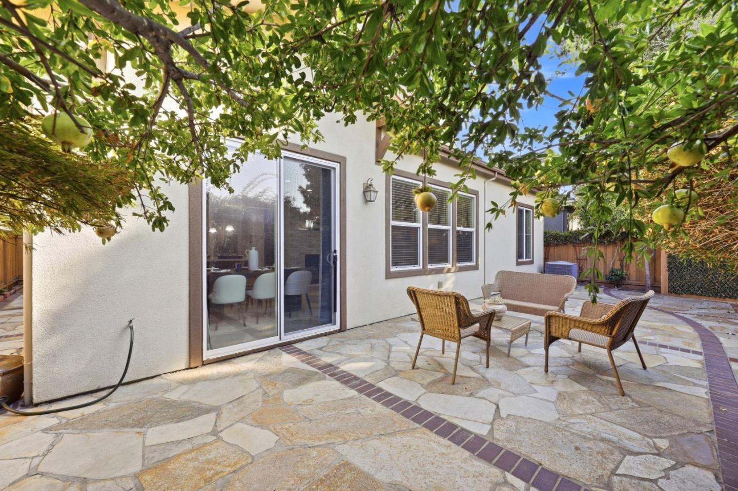 Stone patio with wicker seating beside sliding glass doors, shaded by a leafy fruit tree.