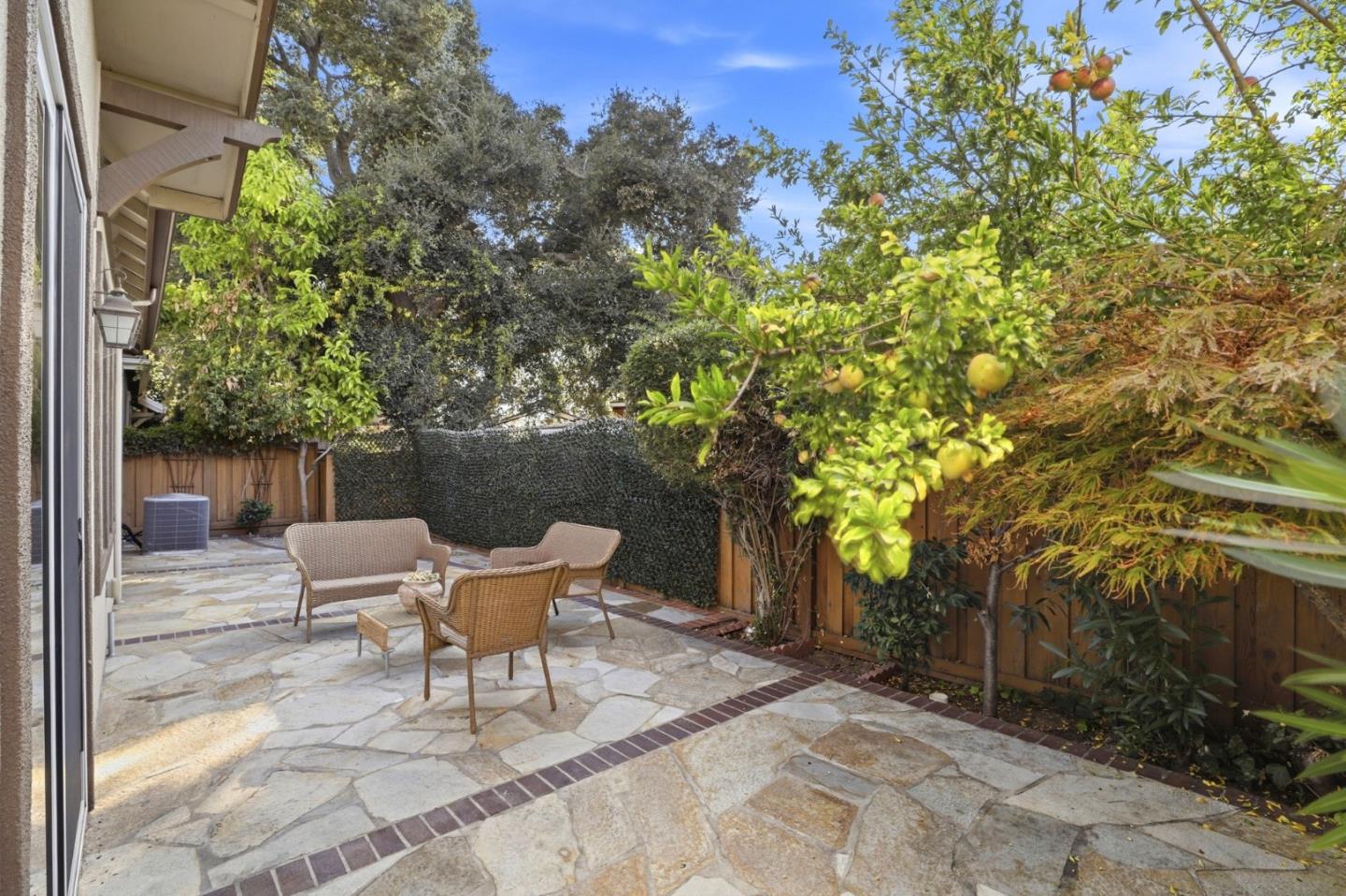 Stone-paved backyard patio with wicker seating beneath fruit trees and leafy fencing.