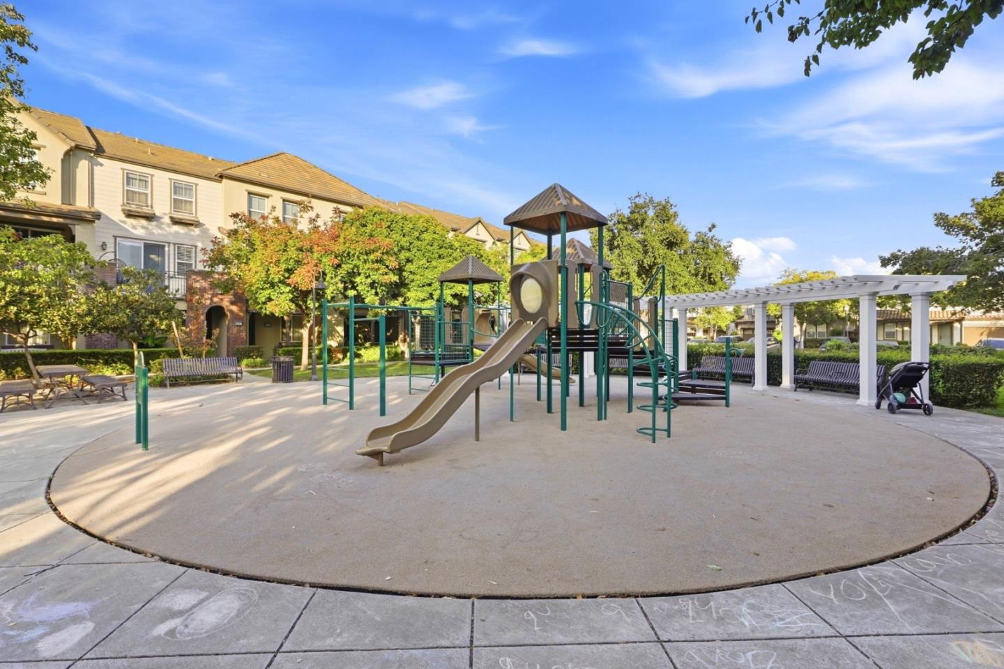Community playground with a large play structure, slides, and a white pergola beside residential buildings.