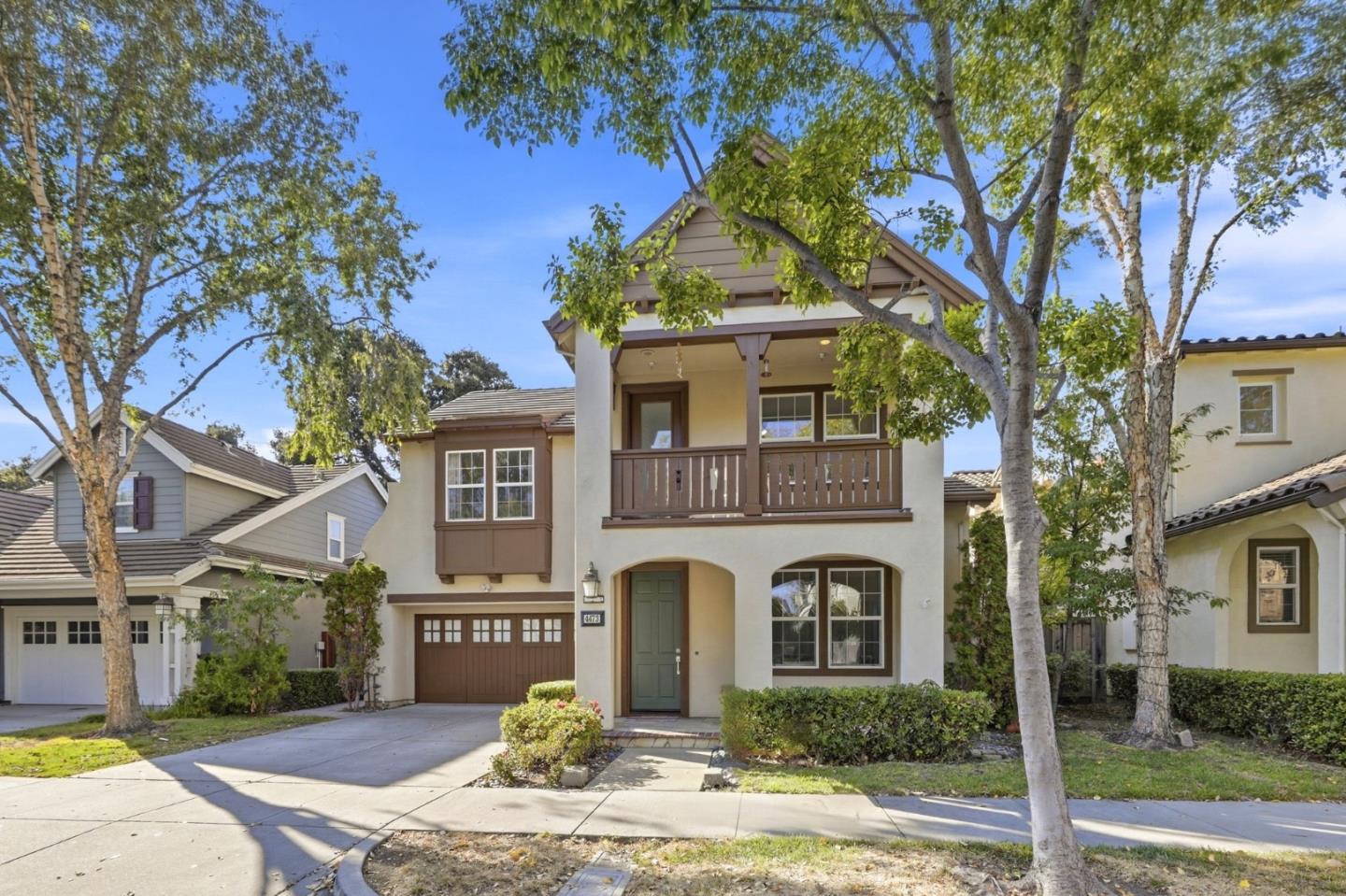 Two-story beige home with a brown garage, upper balcony, and tree-shaded front yard.