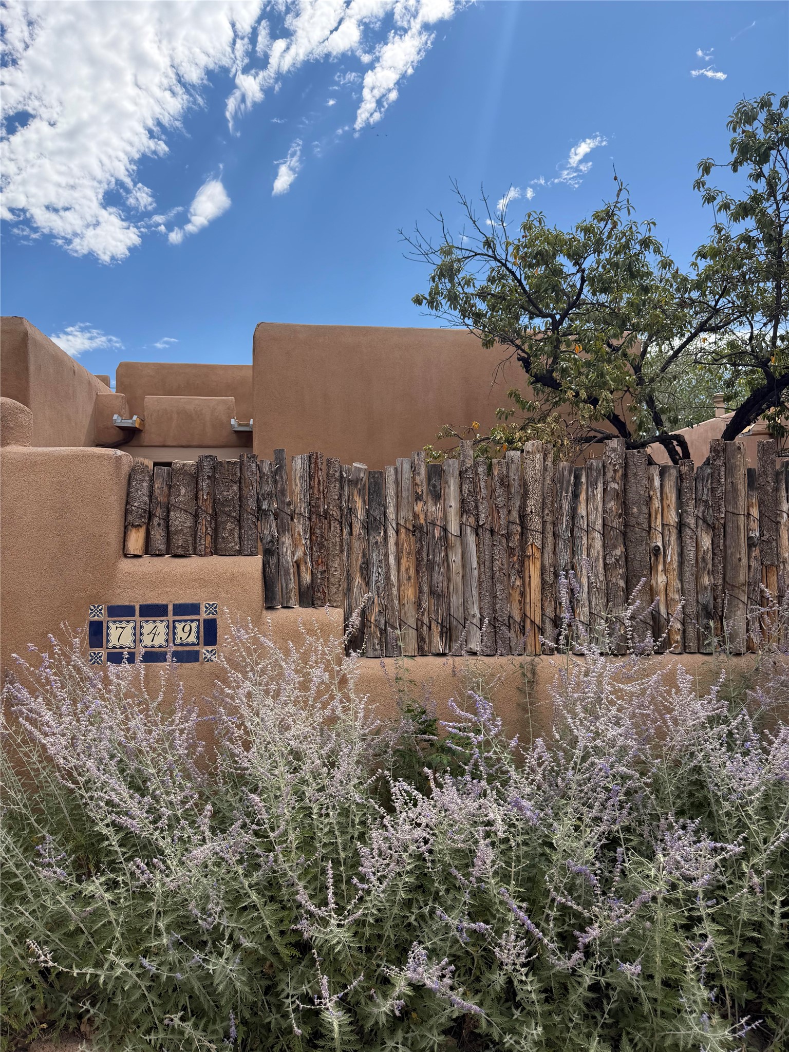 Adobe home behind a rustic log fence, with a tile address marker reading 749 and purple flowers.