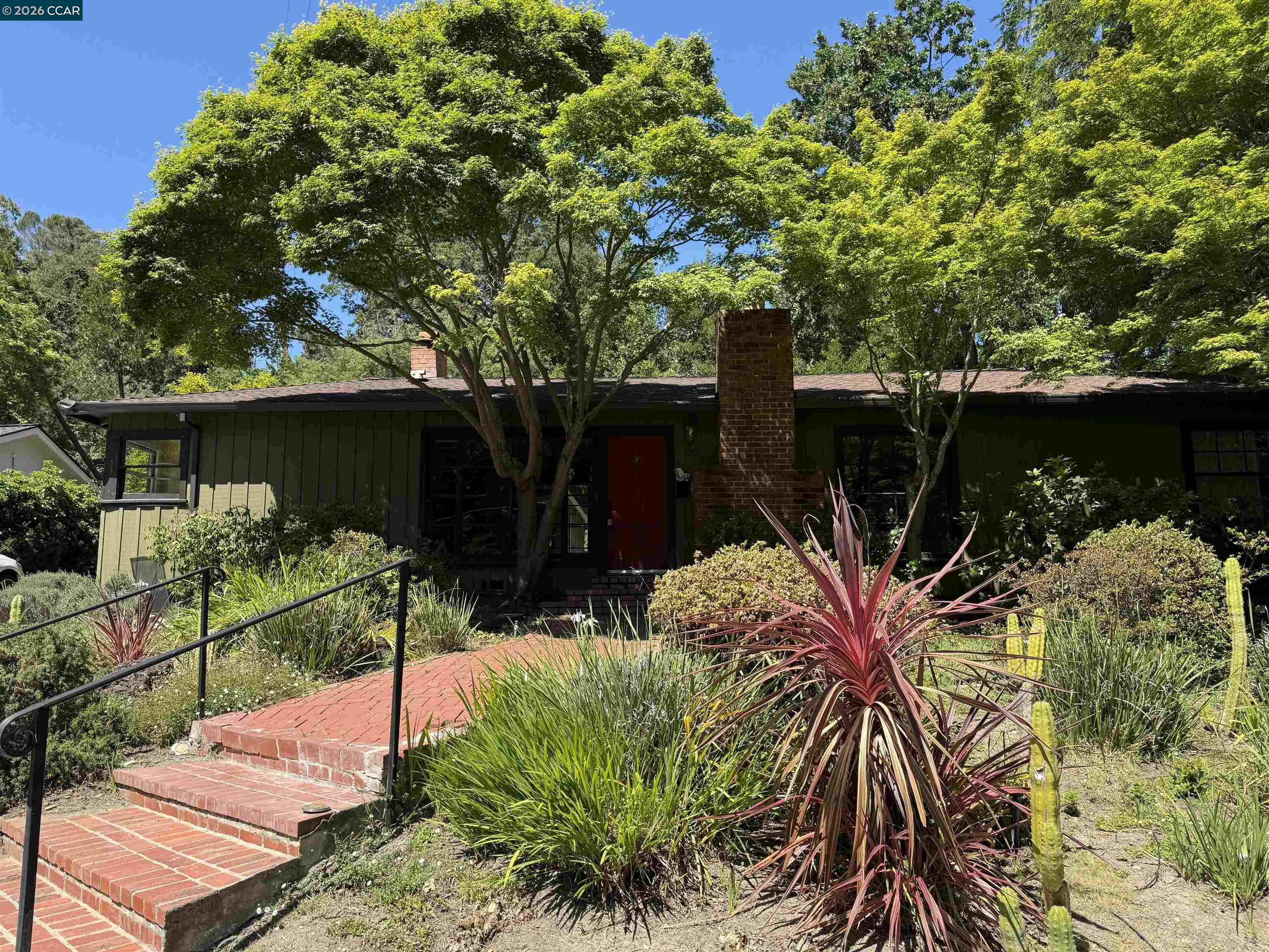 Single-story green home shaded by mature trees, with a brick chimney and landscaped brick entrance path.