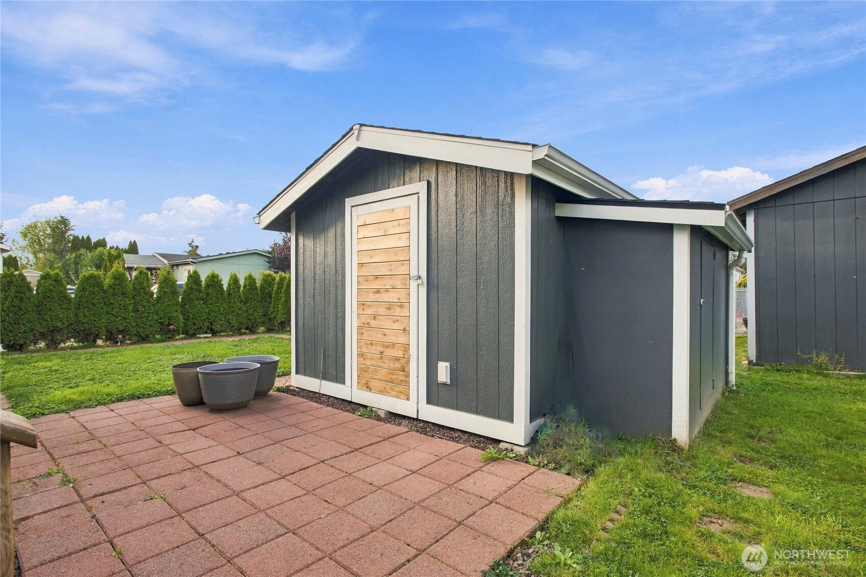 Dark gray backyard shed with a wooden door beside a brick patio and lawn.
