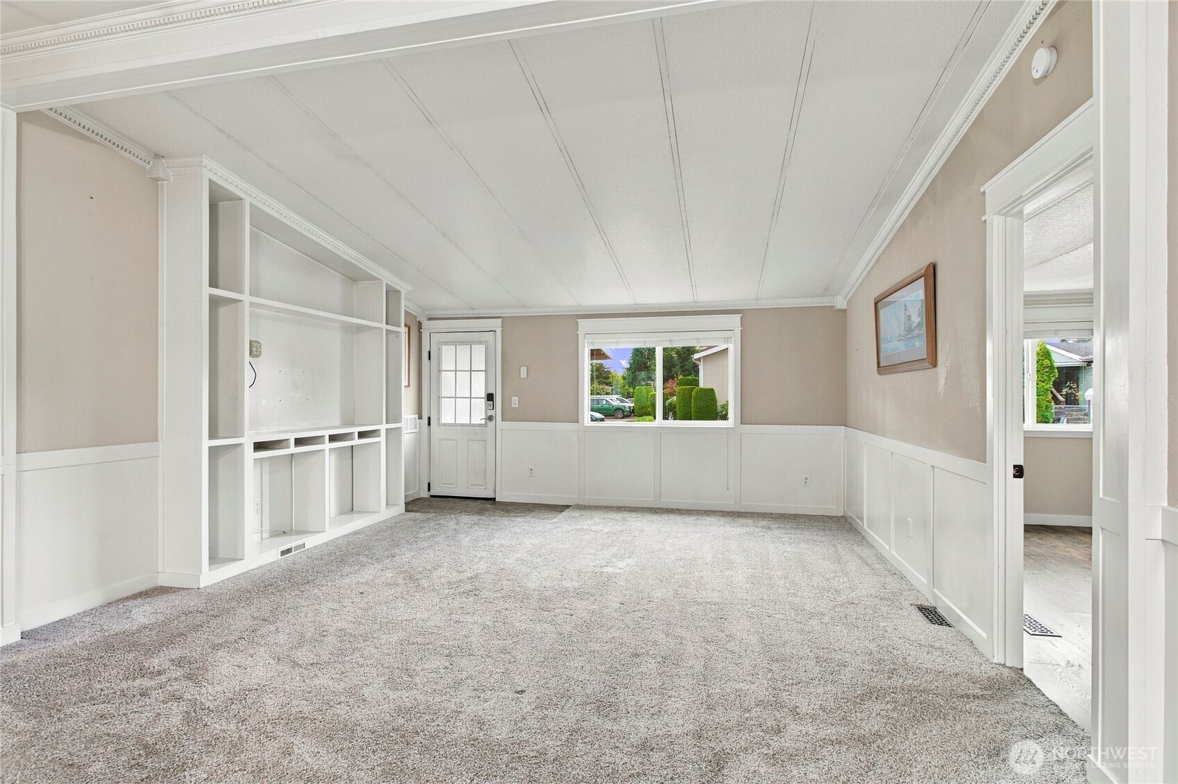 Carpeted living room with built-in shelving, paneled walls, and a window overlooking greenery.