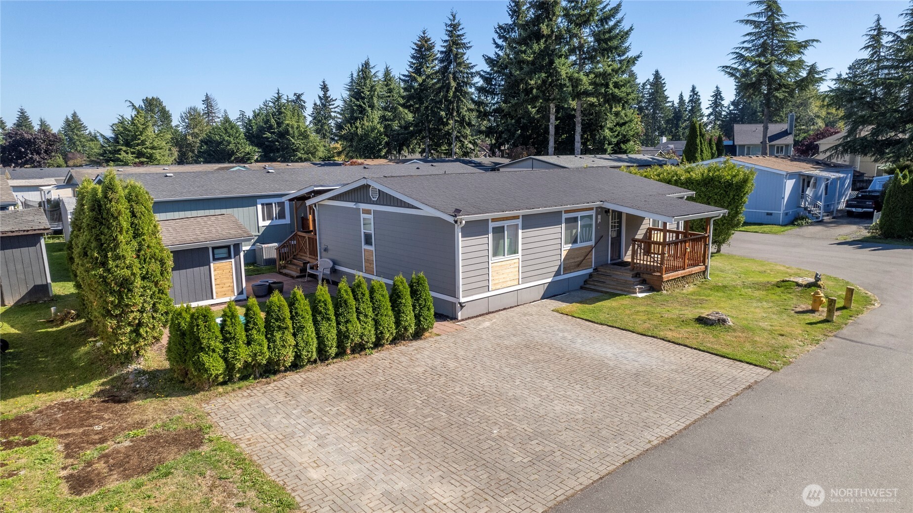 Gray manufactured home with a broad brick driveway, wooden porch, and detached shed.