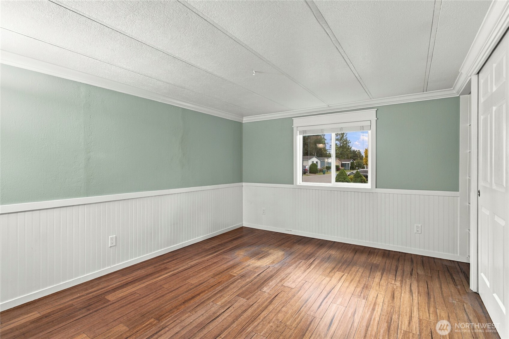 Empty bedroom with hardwood floors, mint-green walls, white wainscoting, and a window overlooking the neighborhood.