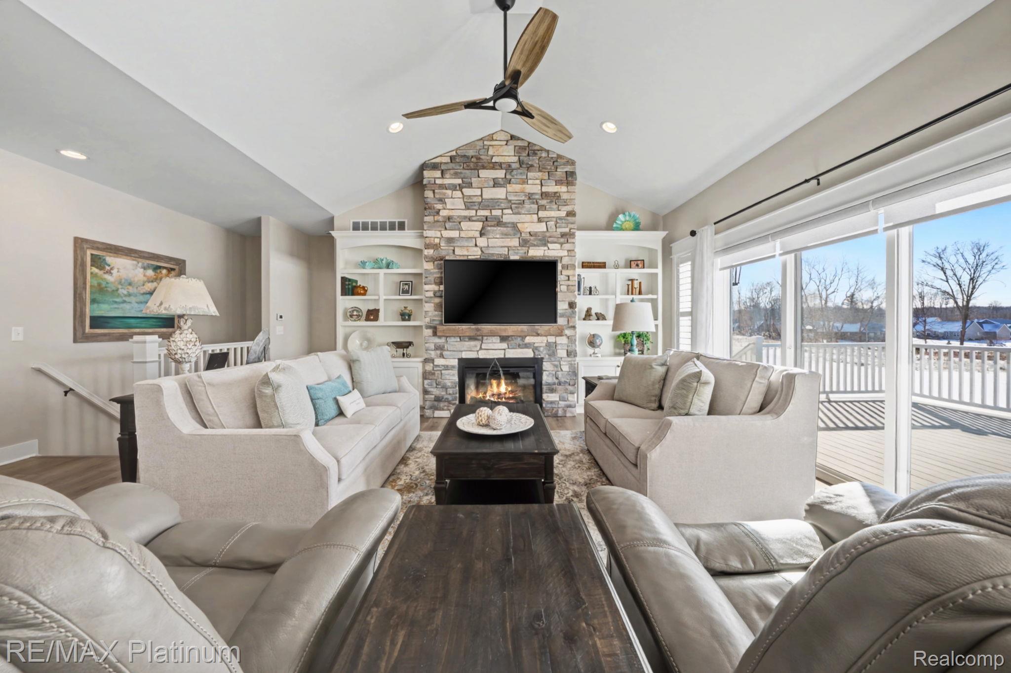 Living room with a vaulted ceiling, stone fireplace, built-in shelves, and windows overlooking a snowy deck.