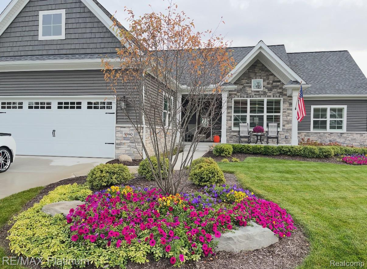 Gray-sided home with stone accents, an American flag, and colorful flower beds in front.