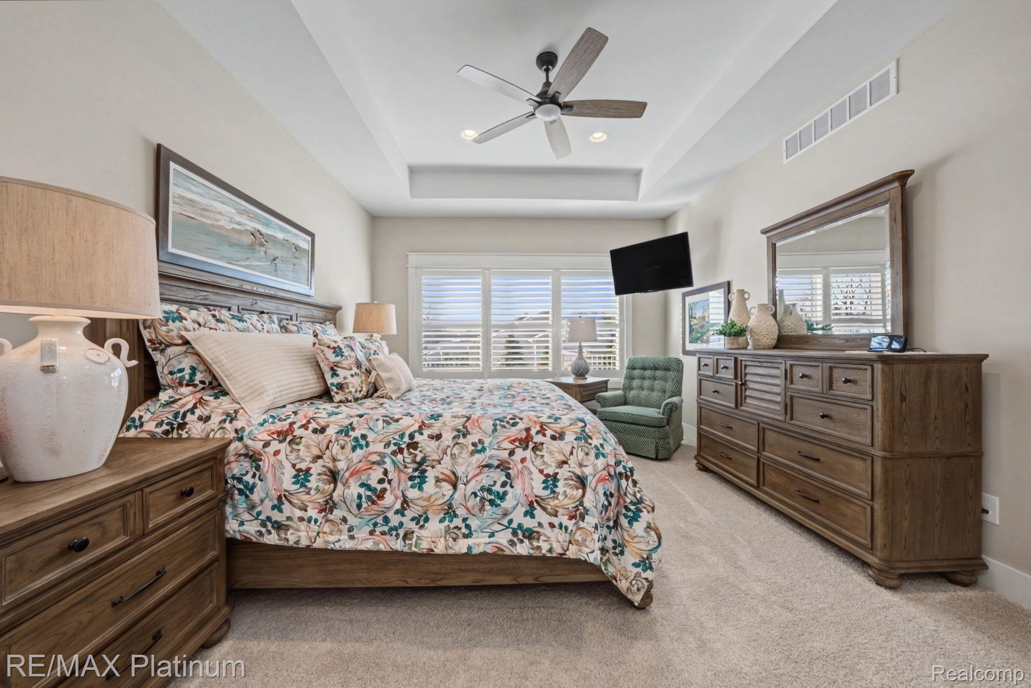 Furnished bedroom with floral bedding, dark wood furniture, plantation shutters, and a tray ceiling.